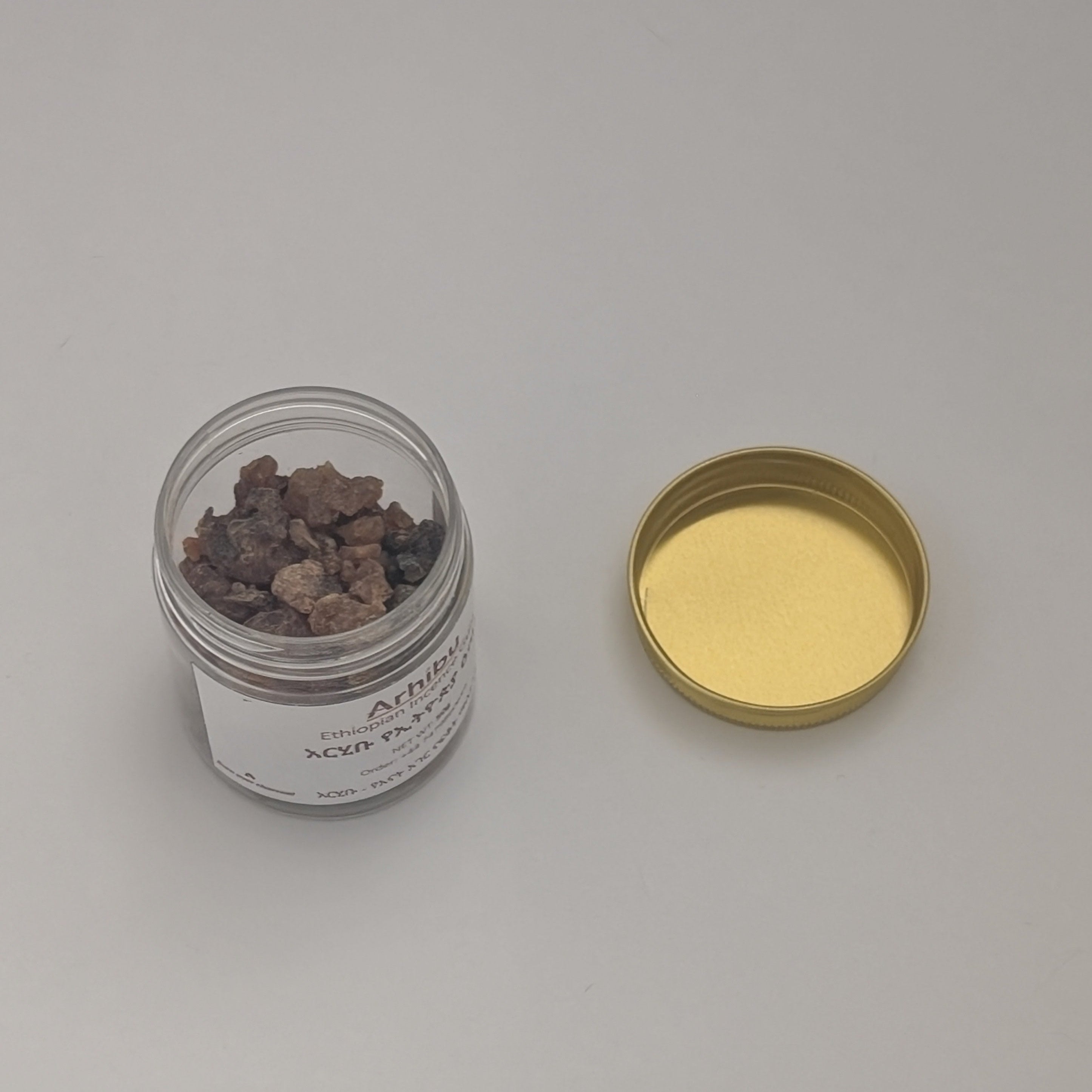 A small clear jar containing brown Kerbe incense gum pieces, shown next to a gold metal lid on a plain light gray background.