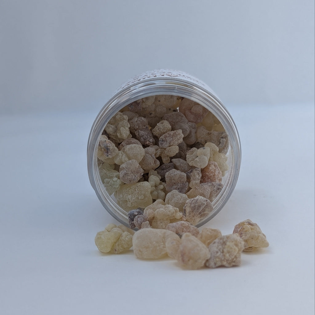 A small clear plastic jar lying on its side with pale beige, translucent rock-like incense gum pieces spilling out onto a white surface against a light gray background.
