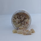 A small clear plastic jar lying on its side with pale beige, translucent rock-like incense gum pieces spilling out onto a white surface against a light gray background.