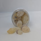 A clear cylindrical jar tipped on its side with pale yellow translucent crystals spilling out onto a white surface. The crystals appear rough and semi-translucent, and a white label is visible on the jar.