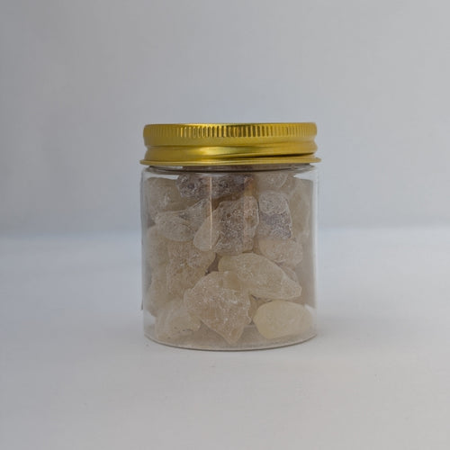 A small clear plastic jar filled with rough, translucent incense gum tears, sealed with a metallic gold screw-top lid, set against a plain light grey background.